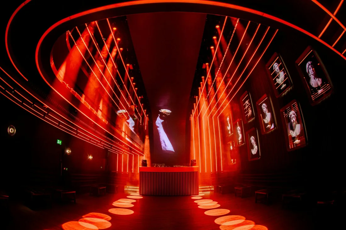 77 empty red-lit club room with DJ booth, neon strips and framed busts