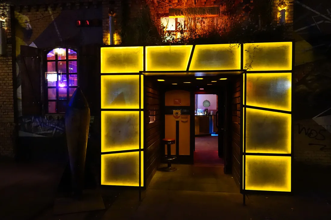 Crack Bellmer lit yellow-panel entrance at night between brick buildings