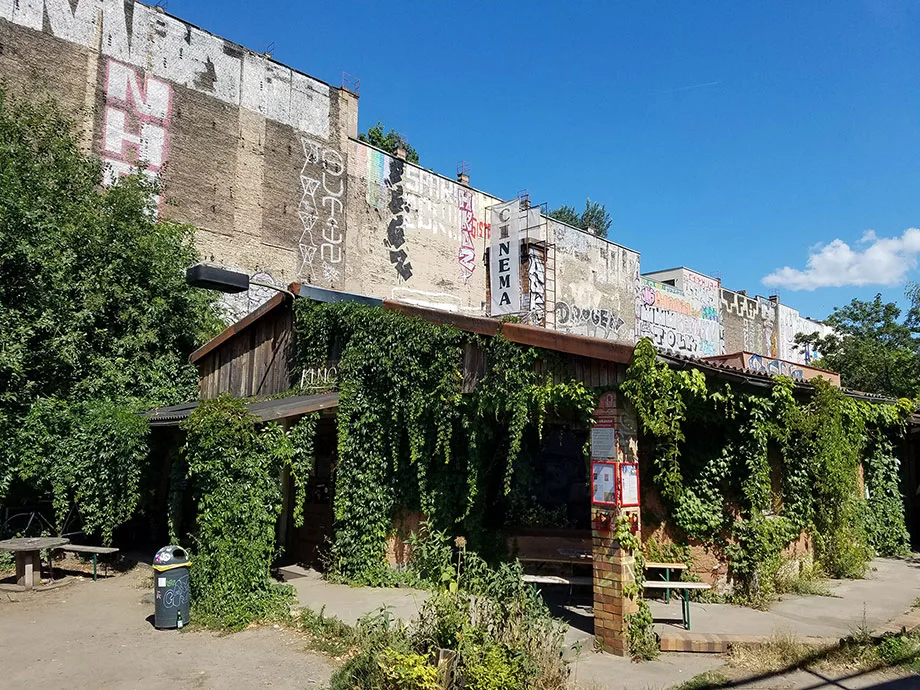 Neue Zukunft ivy-covered wooden cinema hut under graffitied firewalls, daytime